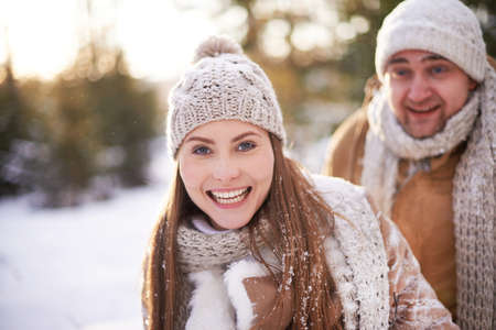 Cute girl in winterwear looking at camera on background of her boyfriend in winter parkの写真素材