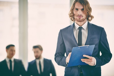Serious young businessman with clipboard looking at cameraの写真素材