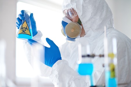 Scientist in protective overall, respirator and gloves holding tube with toxic substance
