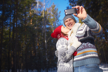 Amorous girl kissing her boyfriend while he making their selfie in natural environmentの写真素材