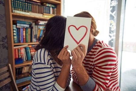 Flirty couple sitting in library behind paper with red heartの写真素材
