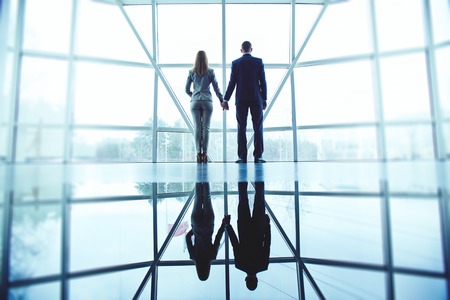 Young man and woman in formalwear holding by hands while standing by office windowの写真素材