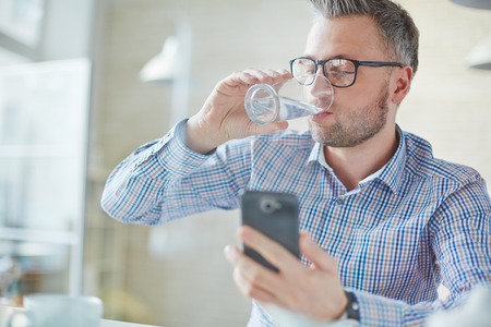 Businessman in casualwear drinking water while reading sms or dialing number on cellphoneの写真素材
