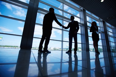 Male office workers standing by the window and handshaking with their colleague near byの写真素材
