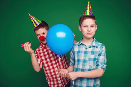 Sly boy in birthday cap going to burst balloon of his brotherの写真素材