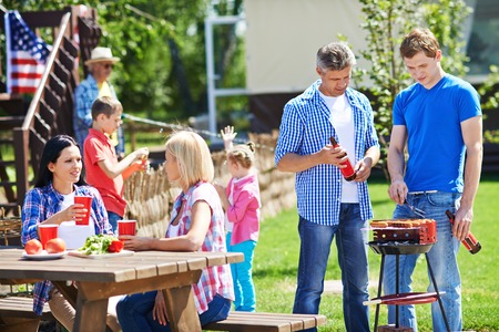 Two men frying sausages on grill outdoors with young women talking by table near byの写真素材