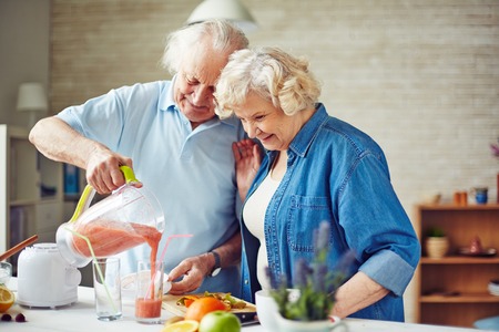 Senior man pouring fruit smoothie into glass with his wife near byの写真素材