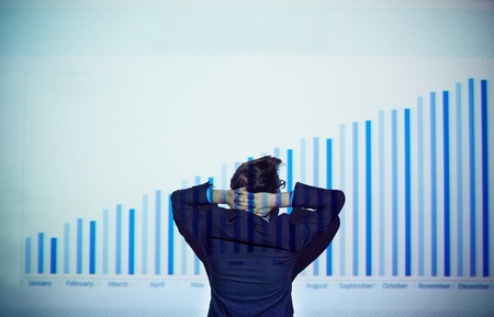 Rear view of businessman with his hands behind head looking at chart on the wallの写真素材