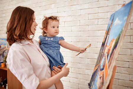 Young mother and her cute little daughter with paintbrushes looking at paintingの写真素材