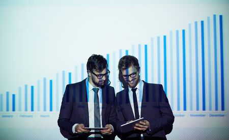 Two colleagues with touchpads networking by chart on the wallの写真素材