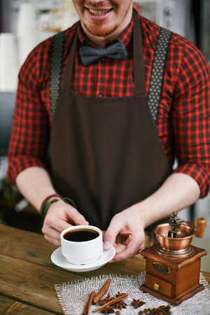 Smiling barista holding cup of black coffee with coffee grinder near byの写真素材