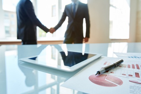 Business objects at workplace with handshaking partners on backgroundの写真素材