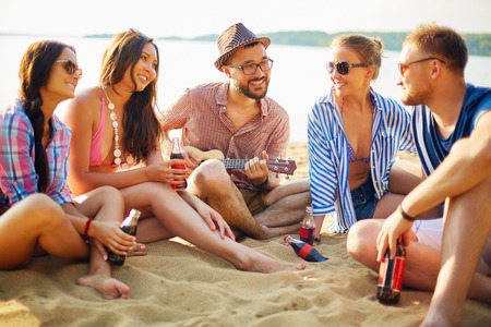 Happy friends with drinks sitting on sand by waterの写真素材