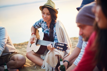 Pretty girl with guitar sitting on sandy beach among her friendsの写真素材