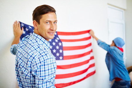 Smiling man looking at camera on background of his son hanging USA flag on wallの写真素材