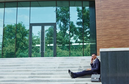 Happy young man in suit speaking on cellphone and typing in urban environment