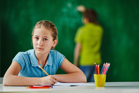Serious pupil looking at camera on background of schoolboy standing by blackboardの写真素材