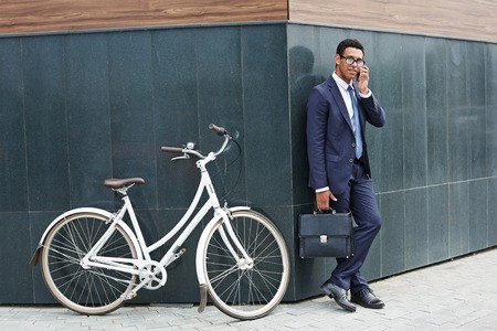 Modern agent speaking on the phone while standing by corner of modern building with bicycle near byの写真素材