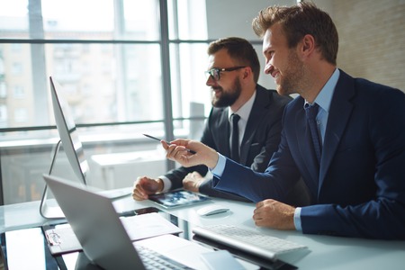 Confident businessman consulting his colleague and pointing at computer monitor at meetingの写真素材