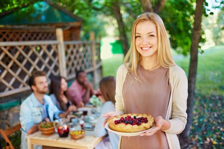 Happy young woman with berry pie looking at camera on background of her friends sitting by Thanksgiving tableの写真素材