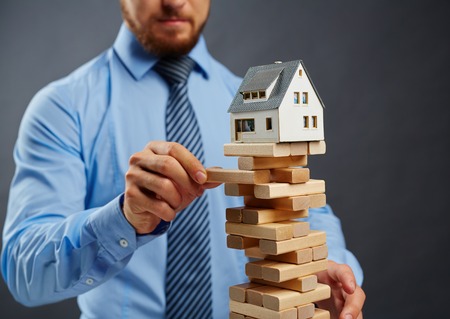 Businessman taking out wooden block from high construction with toy house on its topの写真素材