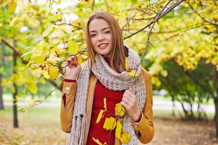 Happy girl in leather jacket, red pullover and knitted scarf looking at camera in parkの写真素材