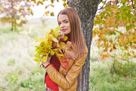 Stylish girl with bunch of foliage standing by tree trunk and looking at cameraの写真素材