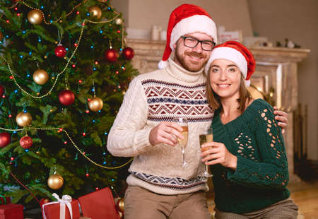 Happy couple in Santa caps holding flutes with champagne by Christmas treeの写真素材