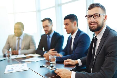 Happy businessman looking at camera at conference with colleagues on background