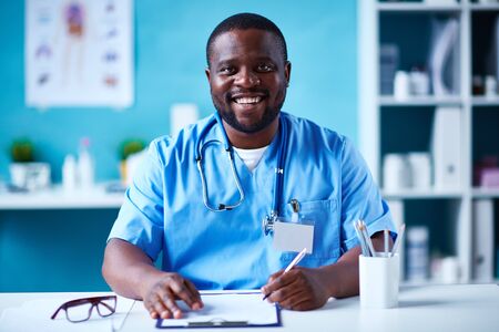 Happy young doctor in uniform looking at camera while making notesの写真素材