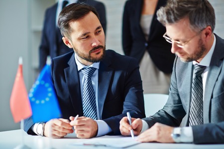 Serious employee looking at business partner signing contract with his colleagues on backgroundの写真素材