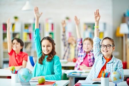 Happy pupils sitting by desks and raising hands at lessonの写真素材