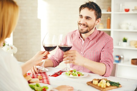 Happy young man with glass of red wine toasting with his wife during romantic dinnerの写真素材