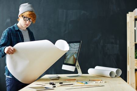 Young serious architect looking at blueprint by workplace