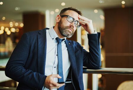 Pensive employee with cellphone leaning on railings in business centerの写真素材