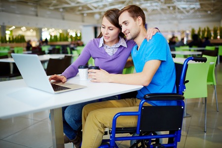Amorous girl embracing her boyfriend in wheelchair while networking in cafeの写真素材