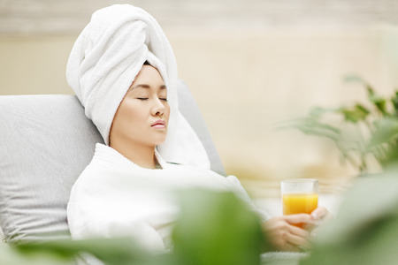 Young Asian woman with towel on her head and fruit juice in her hands relaxing in spa salonの写真素材
