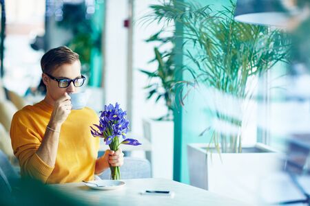 Young man with flowers drinking coffee at cafeの写真素材