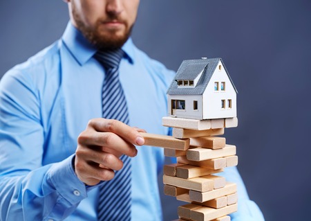 Modern businessman taking out wooden block from tower with toy house on its topの写真素材