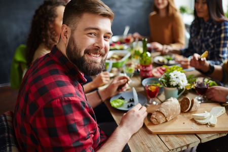 Young man sitting at dinner table with his friends and smiling at cameraの写真素材