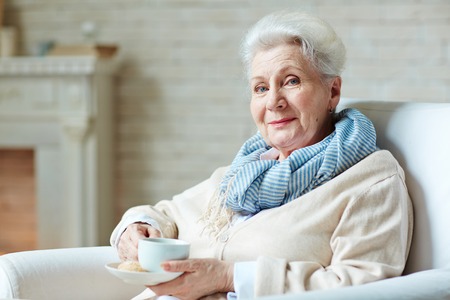 Senior female sitting in arm-chair with cup of tea and looking at cameraの写真素材