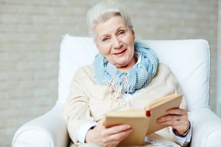 Happy senior female sitting in arm-chair with open book and looking at cameraの写真素材