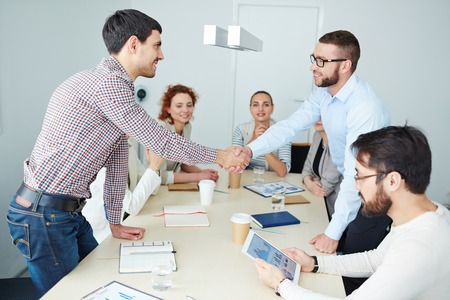Two young managers handshaking over table in office among his colleaguesの写真素材