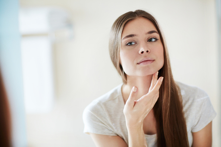Young woman looking at mirror at home and applying cream on her faceの写真素材