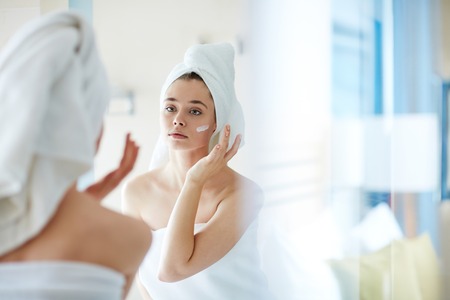 Young woman applying foundation or moisturizer on her face in front of mirrorの写真素材