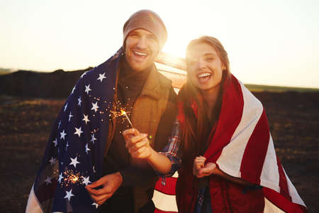 Happy young patriots with American flag and bengal lights looking at cameraの写真素材