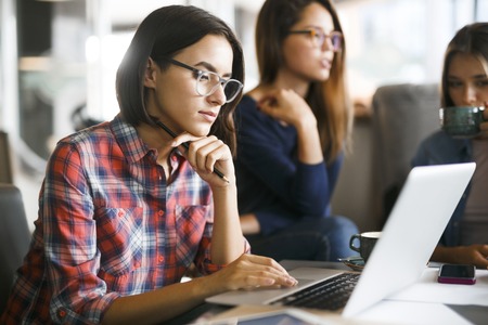 Serious student with laptop networking with her friends on backgroundの写真素材