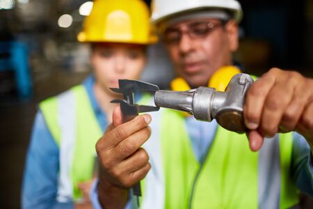 Engineer with metallic machine part measuring and showing it to his assistant