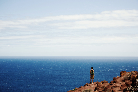 Young man standing on cliff and looking at blue oceanの写真素材