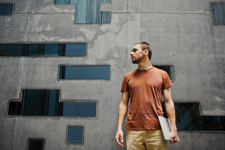 Serious young man with laptop standing against wall of modern buildingの写真素材
